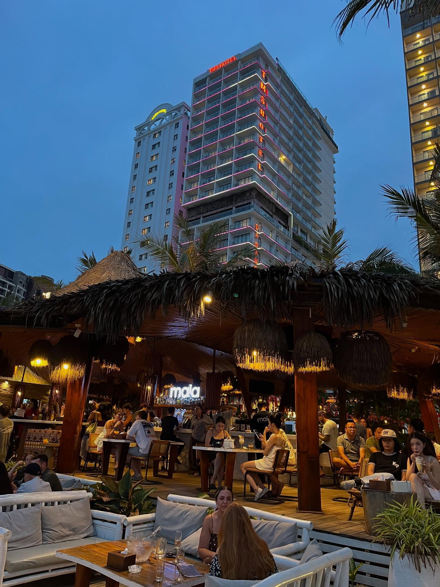 Maia Beach Bar's beachfront deck packed at blue hour, the M Hotel tower lit behind