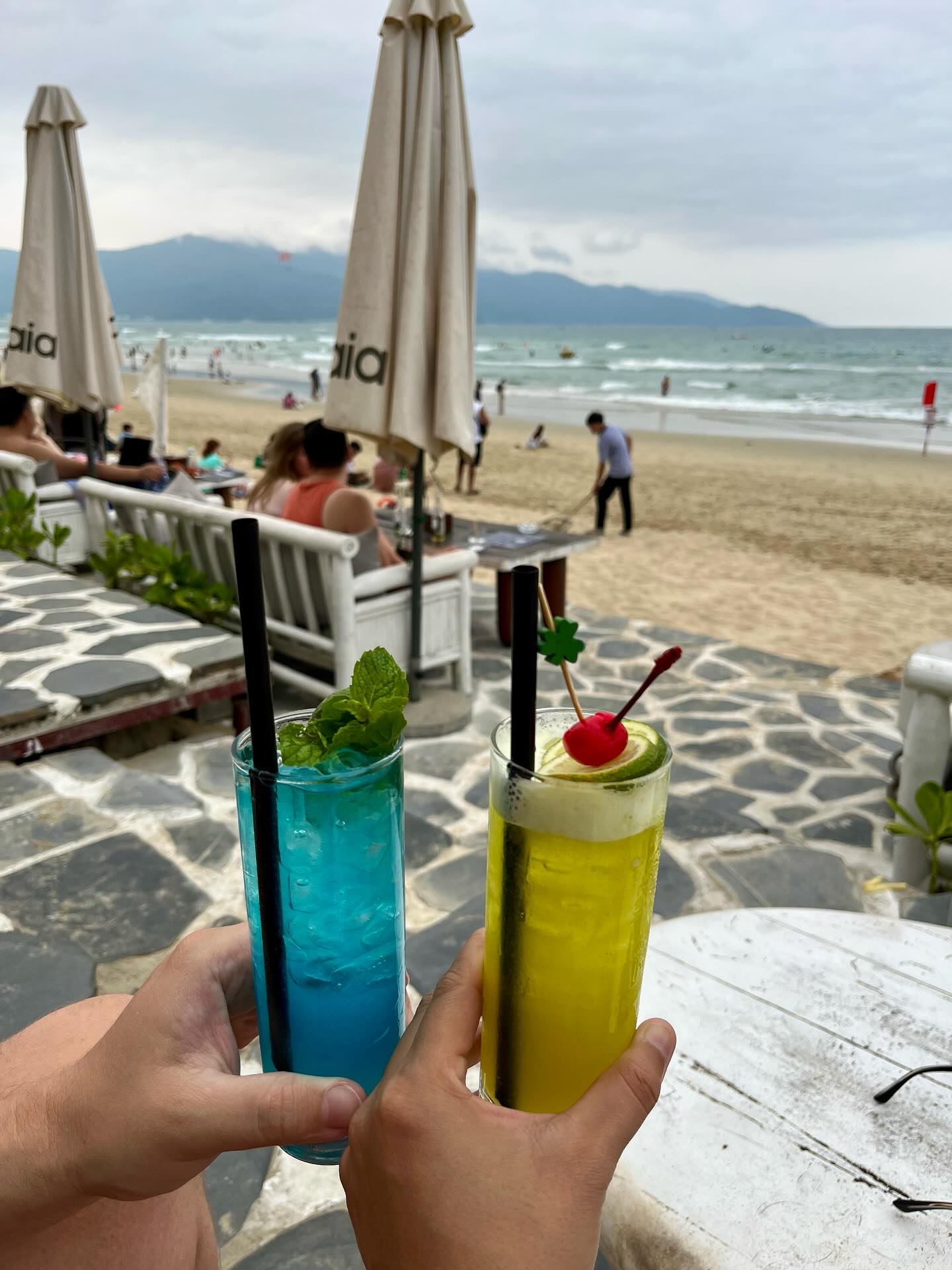 Two cocktails held up over the sand, Son Tra and the Marble Mountains behind