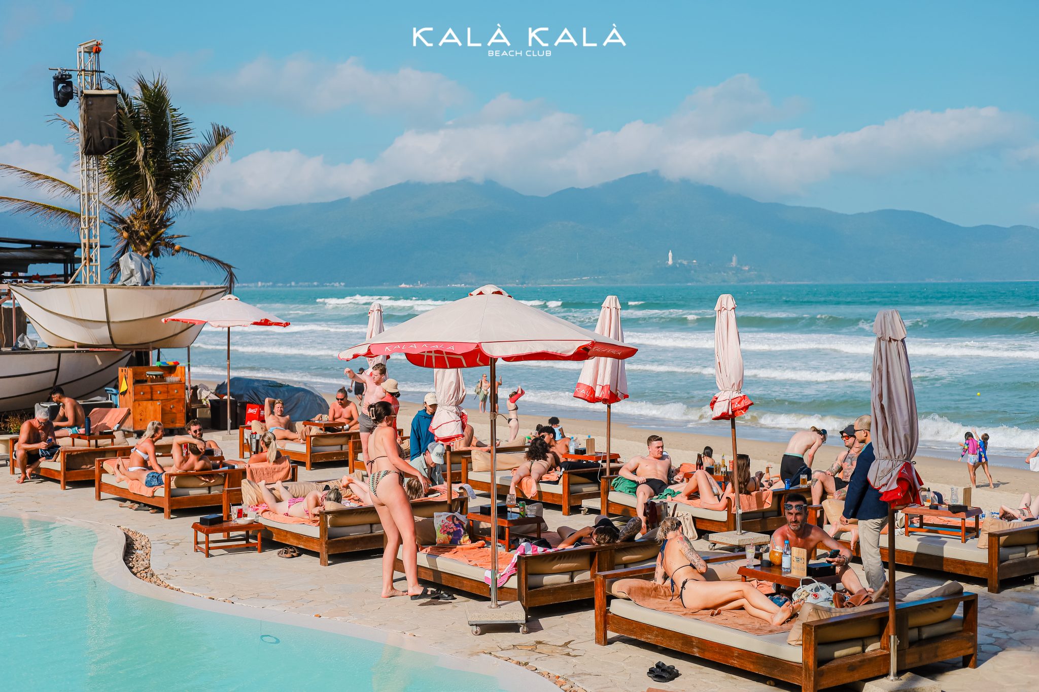 Packed daybeds and red parasols by the pool with ocean and mountains beyond
